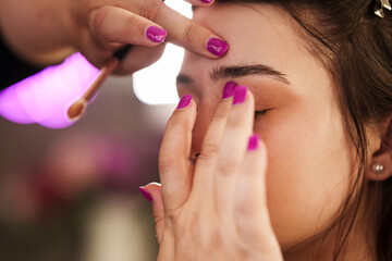 Professional beautician applying makeup on beautiful young woman's face. Professional make up beauty studio work with gorgeous female model.