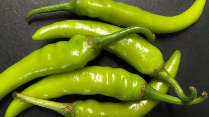 Close-up photo of fresh green chili peppers arranged on a dark surface. The vibrant color and glossy texture highlight the freshness of the peppers, perfect for use in food, spice, and cooking.