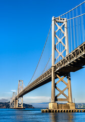 A detailed perspective of the iconic San Francisco-Oakland Bay Bridge's tower and cables, stretching over the bay on a bright day with a clear blue sky. Symbolizes engineering and urban travel