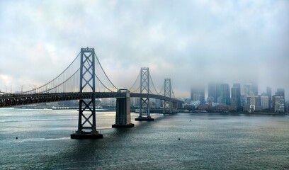 San Francisco-Oakland Bay Bridge on a moody, foggy day. The city skyline is partially hidden by low clouds, and the bay water is calm in the foreground. Represents travel and urban atmosphere