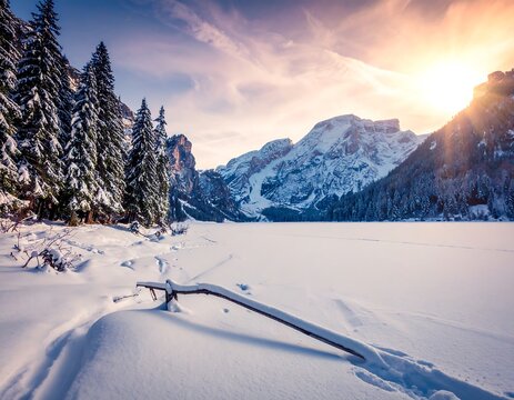 A scenic winter landscape featuring a snow-covered lake, evergreen trees, and majestic mountains under a sunlit sky