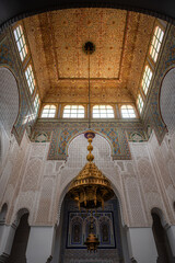 Upward view of the ornate ceiling and richly decorated walls of the Mausoleum of Moulay Ismail in Meknes, Morocco, showcasing intricate geometric patterns, stucco carvings, and colorful tilework.

