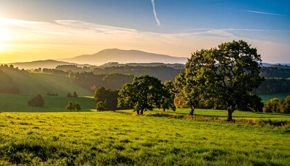 A scenic rural landscape at golden hour, featuring rolling hills, lush green meadows, mature trees, and a mountain range in the distance