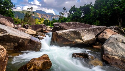 A serene river flows over and around large, moss-covered boulders, surrounded by lush green trees and distant mountains under a cloudy sky