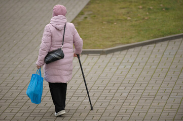 An elderly woman with a cane carries heavy bags along the street
