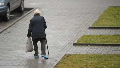 A lonely gray-haired homeless woman with a cane and a bag in her hand walks down the street