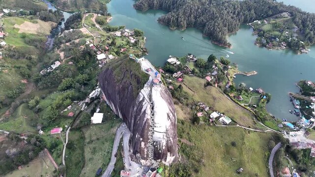 A dynamic aerial pulls back from the top of the Guatap&eacute; rock and pans upward, revealing the entire massive monolith, surrounding lake, islands, and landscape under a cloudy sky