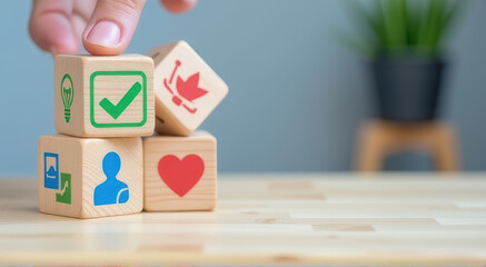 Wooden blocks with colorful icons including lightbulb, check mark, leaf, person, and heart are stacked on wooden table. hand is adjusting one block. blurred plant is in background
