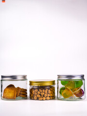 Three glass jars filled with delicious snacks cookies peanuts colorful jelly candies beautifully arranged on clean white surface