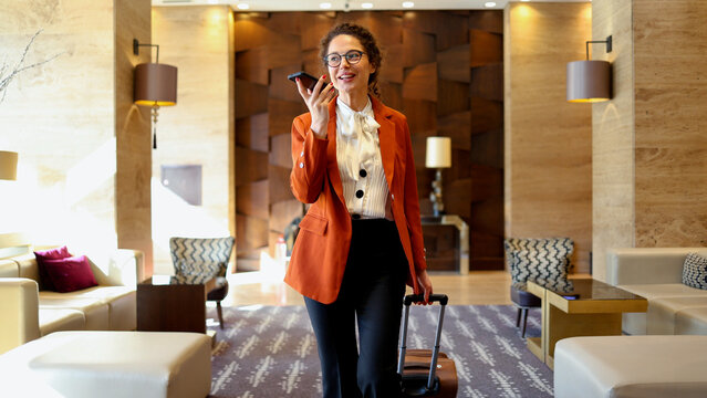 Businesswoman walking in modern hotel lobby, speaking on smartphone with suitcase, professional travel and communication concept