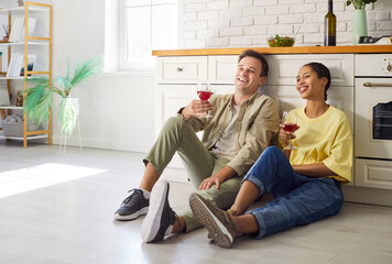 Couple relax at home kitchen, sharing wine. Smiling partners sit on the floor and toast, enjoying love, family time, and bright lifestyle decor nearby. Cozy day together, sharing happiness.