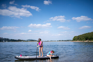 Girl in life jacket and baseball cap paddling on a SUP board, standing at the front with her sister behind, bright blue sky with clouds, forest and shore in the background, copy space