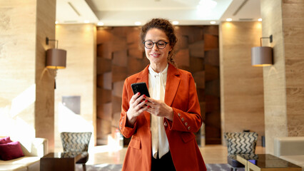 Professional businesswoman in eyeglasses browsing smartphone, standing in luxury hotel lobby, smiling and communicating