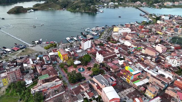 Aerial high-angle view of the vibrant town center and waterfront malec&oacute;n of Guatap&eacute;, Colombia, showcasing the main square park, the marina with boats, and colorful architecture