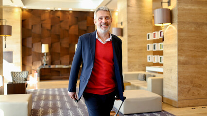 Mature businessman traveling, entering a modern hotel lobby, pulling a trolley bag, and smiling confidently