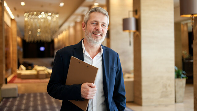 Professional businessman smiling, holding clipboard in luxury hotel lobby, senior corporate leader hospitality