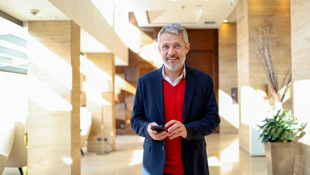 Mature businessman smiling, holding smartphone in modern hotel lobby, connecting and communicating - Powered by Adobe