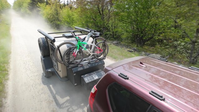 A family enjoys a camping trip as they travel down a dusty road in a teardrop trailer, bicycles secured on the rear, surrounded by lush green trees.