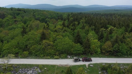 Naklejka premium A family enjoys a camping adventure in a teardrop trailer, parked along a dirt road surrounded by vibrant green trees under a cloudy sky.