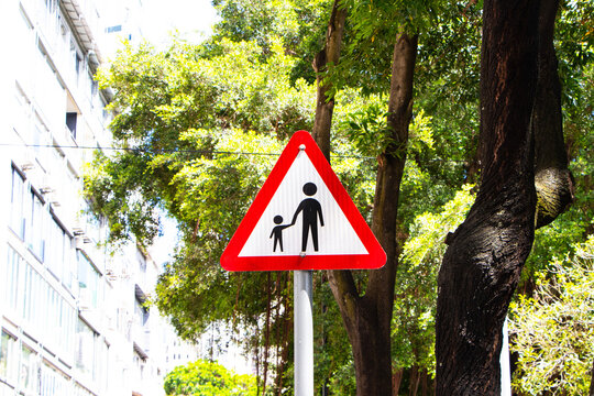 School roadside warning sign students or people crossing and green trees, building city is background. Symbol white triangular aluminum, red post about pedestrian crossing road sign traffic.