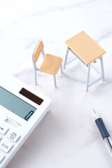 学校の机と椅子、電卓がイメージする、高騰を続けて家計を圧迫する教育費のイメージ
