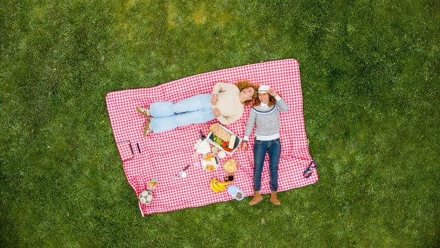 Two women laying on a red picnic blanket with food, enjoying leisure time outdoors