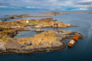 Rusty shipwreck partially submerged in calm coastal waters near a red boathouse on a rocky islet. The Atlantic Ocean Road stretches across Norways wild and rugged seascape