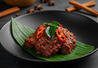 Rendang, a rich and flavorful Indonesian meat dish, is presented on a black plate with a banana leaf garnish, accented with chili slices and curry leaves.