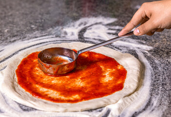 Closeup hand of chef baker making pizza at kitchen. Chef pours tomato sauce over the rolled dough. Chef hand is spreading pasteurized tomato paste onto a pizza base