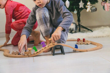 A boy and a girl are playing with a wooden toy train near a Christmas tree at home.