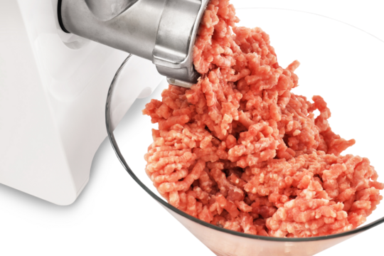 A close-up of fresh, ground meat being extruded from a metal meat grinder and collecting in a clear glass bowl.