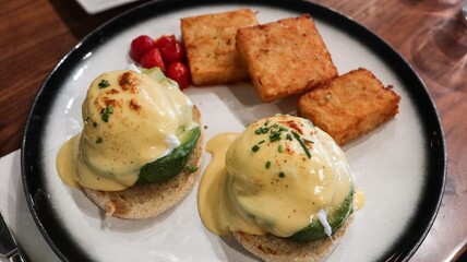 close up of avocado egg Benedict (two poached eggs atop toasted English muffins with creamy hollandaise sauce and sprinkled with  chives, two square golden crispy hash crown and cherry tomato