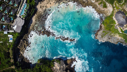 High-angle drone shot capturing the intense turquoise waters of the Blue Lagoon, Nusa Ceningan. Waves crash into the rocky cove, showing the dramatic, vivid colors of the sea.