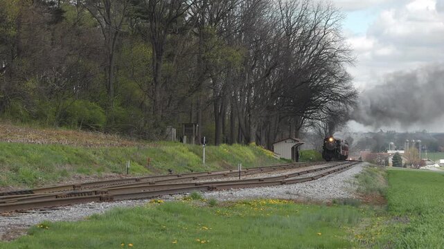 A steam locomotive chugs along the tracks in a scenic countryside, passing a small station. The train emits black smoke against a backdrop of green fields and trees, creating a picturesque view.