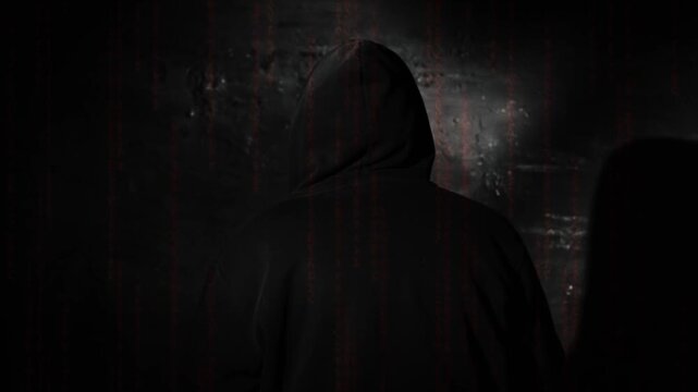 Anonymous hooded hacker sitting with back to camera during tense police interrogation in a dark station room, glowing matrix style code overlay - Powered by Adobe