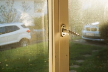 Weißer Fensterrahmen aus Holz mit silbernem Griff und beschlagenem Fensterglas mit Ausblick auf grüne Gartenwiese, parkenden Autos und gegenüberliegendem Wohnhaus bei Sonne am Nachmittag im Herbst