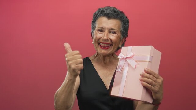 Senior woman with grey hair gives thumbs up while holding gift box against red background, showcasing joy and celebration.
