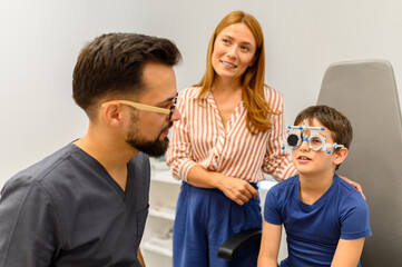 Mother standing beside son wearing phoropter while male doctor examines eyes during routine vision checkup at clinic