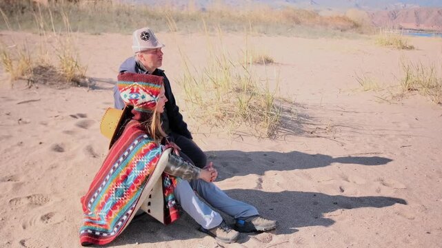A man and woman sit side by side on the sand among coastal dunes. The girl wears a bright patterned poncho and gazes thoughtfully ahead. Peaceful family moment in nature, full of warmth and silence.