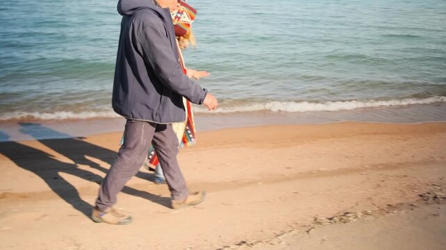 An elderly man and a young woman walk closely side by side along a sandy beach, wrapped in a shared colorful blanket. Peaceful atmosphere, ocean background, generational bond, winter walk.