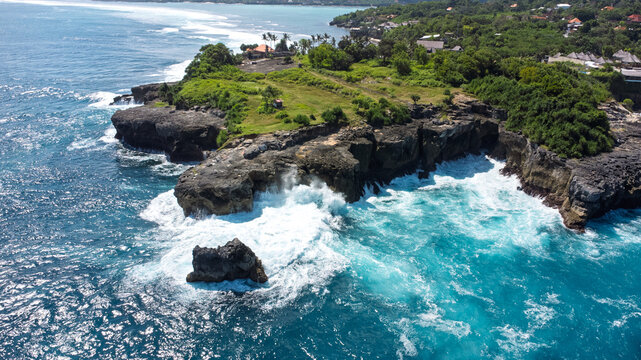 Panoramic aerial view of the green, rugged cliffs above the Blue Lagoon, Nusa Ceningan. Powerful white surf breaks against the rocky shore and the brilliant turquoise water.