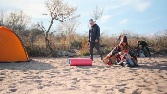Young camper woman wrapped in a blanket sits by a fire pit with a hot drink and smartphone on the seashore. Friends in the background set up a tent and camping equipment.