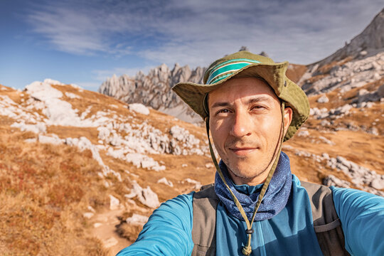 Man wearing a hat taking a selfie while hiking in rocky mountains with dry grass