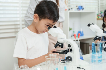 Elementary asian boy exploring science with microscope during classroom experiment while focusing on slide sample and sitting at white table surrounded with lab equipment and glass beakers