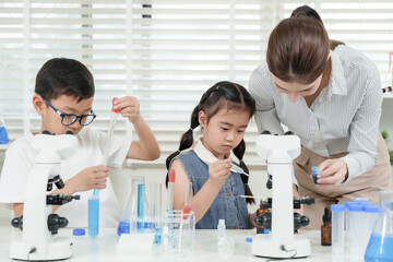 Asian boy using dropper while girl observes and female teacher assists both students during lab experiment in elementary science class colorful tools and glassware arranged neatly
