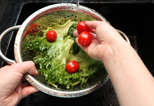 Hands washing fresh lettuce and cherry tomatoes in a metal colander under running water in a black sink. Emphasizing the necessity of hygiene before consumption
