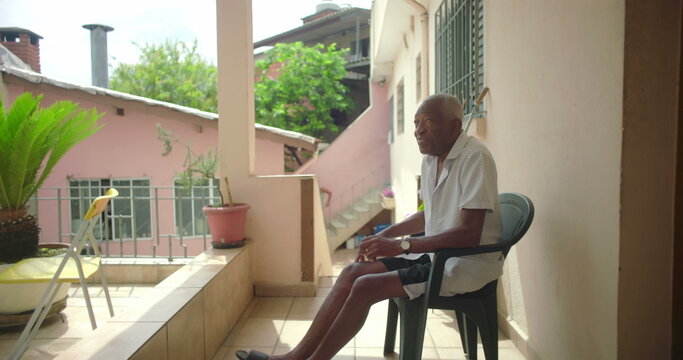 One Elderly Black man seated outdoors on porch, gazing thoughtfully, surrounded by warm tropical architecture, pink walls, serene and contemplative expression