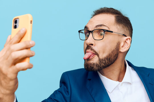 A professional man in business attire poses for a self portrait on an isolated colored backdrop, projecting confident focus and playful emotion while using a smartphone.