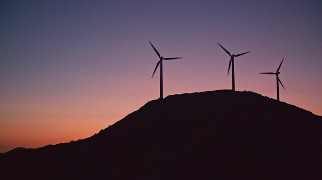 A row of wind turbines silhouetted against a vibrant sunset sky on a rural hillside. The spinning blades highlight clean, renewable wind power and sustainable energy production.