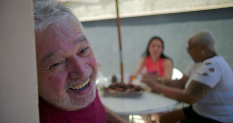 Elderly man smiling joyfully while holding a glass at a backyard barbecue, enjoying food, laughter,...